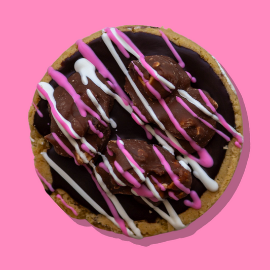 Rocky Road Maxi cookie with chocolate ganache, marshmallows, nuts, and pink and white chocolate drizzle on a pink background.