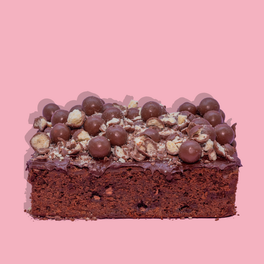 Loaded Brownie topped with chocolate balls and nuts on a pink background.